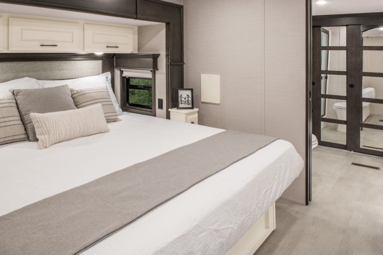 Modern bedroom interior in a luxury RV, featuring a neatly made double bed with white and grey bedding. A small nightstand holds a framed picture, while windows with blinds offer privacy. Glass doors reveal an adjoining bathroom, enhancing the light and minimalistic decor of this mobile haven.