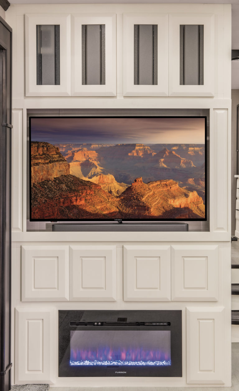 A wall-mounted television in a luxury RV displays a scenic desert canyon landscape. Below, an electric fireplace emits soft blue and purple flames. The setup is surrounded by white paneled cabinetry with glass accents, creating an oasis of elegance on the road.