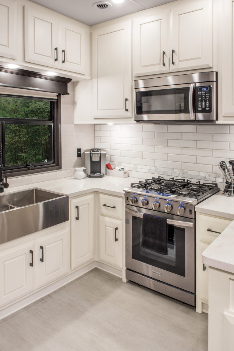 Modern kitchen in a luxury RV features white cabinets, stainless steel appliances, and a subway tile backsplash. Includes a farmhouse sink, gas stove with oven, microwave, and coffee maker. A dish towel hangs neatly on the oven handle below the stove.