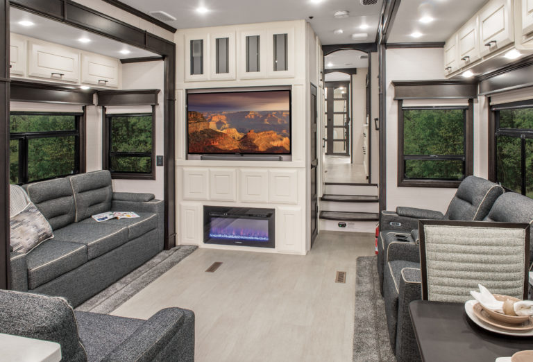 Explore the interior of this luxurious RV, where modern elegance meets comfort. Relax on gray sofas, enjoy a large TV above an electric fireplace, and dine in a chic kitchen nook. Windows flood the space with natural light and views of greenery—your perfect escape among luxury RVs for sale.