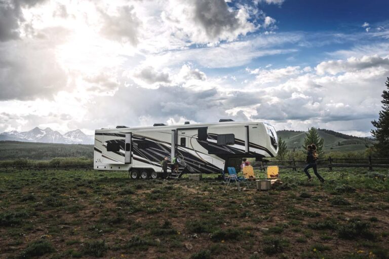 A large luxury RV is parked in an open grassy area, surrounded by mountains and a partly cloudy sky. Two people sit on camping chairs by a picnic table, while another person stands nearby, capturing the scene with a camera.