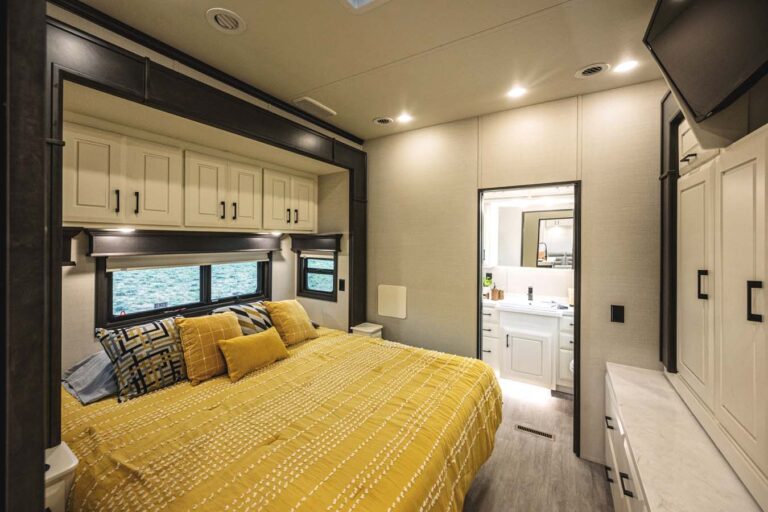 A cozy and modern bedroom in a luxury RV features a large bed with a yellow patterned duvet and pillows. Cabinets are above the bed, with a window on the side. Through an open doorway, an adjacent bathroom with a sink and mirror completes this exquisite luxury 5th wheel RV setup.