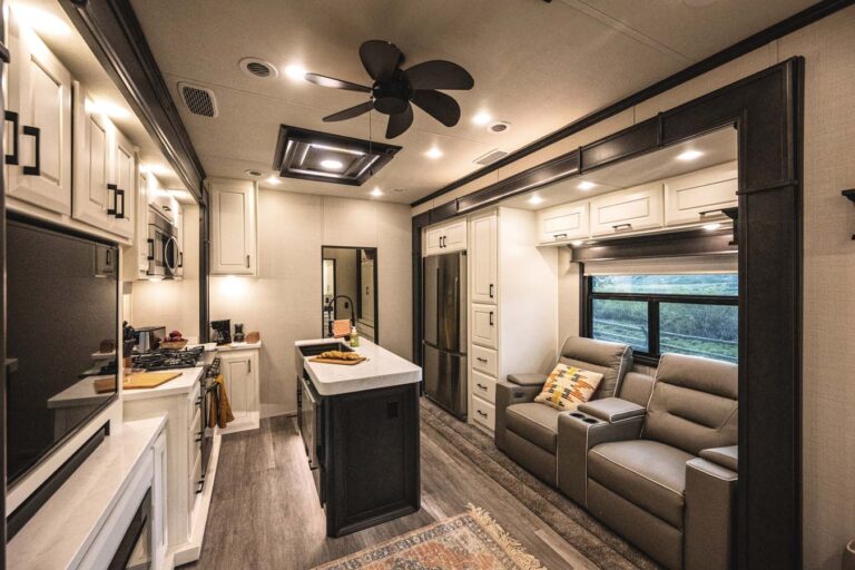 The interior of this luxury RV boasts a kitchen with white cabinetry, a stove, and a refrigerator. Opposite are two recliners beside a sizable window. The ceiling is adorned with recessed lighting and a fan, while a decorative rug enhances the wooden floor’s elegance.