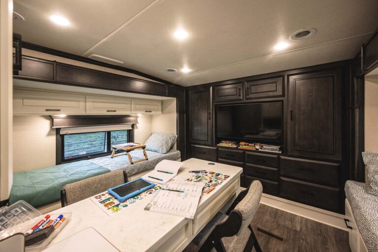 The interior of this luxury RV features a sleek, modern design. A light-colored dining table laden with games and papers is surrounded by cushioned chairs. A bed with a green cover sits beside a window, while dark wood cabinets and a TV adorn the opposite wall, exuding comfort and style.