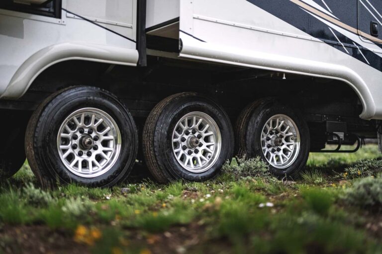 Close-up of three wheels of a luxury RV with shiny metal rims. The RV is parked on grass speckled with small wildflowers, and the sleek exterior of this high-end vehicle is partially visible above the wheels.