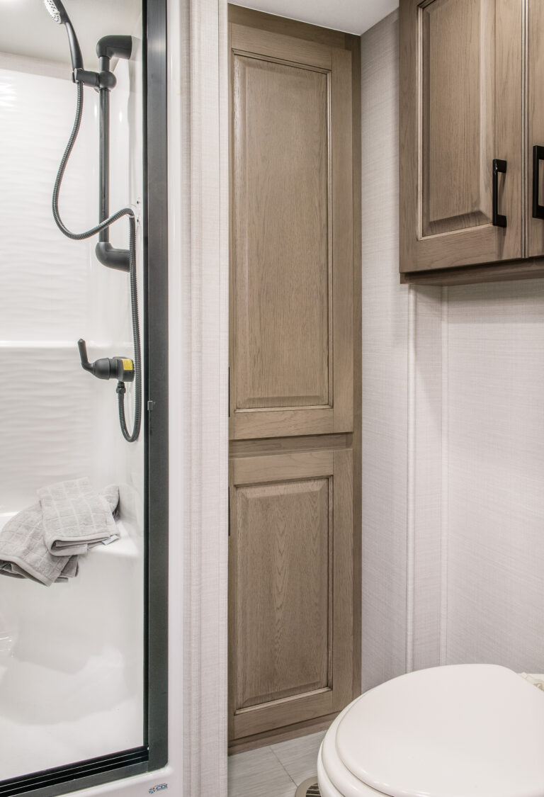 Compact bathroom in a luxury RV with a corner shower featuring a black showerhead and glass door. A wooden cabinet is next to it for storage, while a white toilet is partially visible in the foreground. Neutral tones create a clean, modern look.