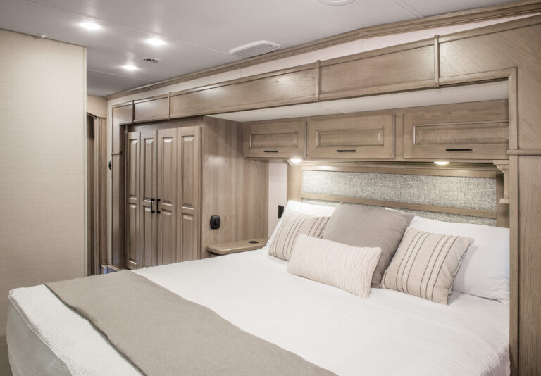 Experience the epitome of comfort in this modern RV bedroom, where luxury RV design meets functionality. A spacious bed with plush white and beige linens invites relaxation, while elegant wooden cabinetry and soft lighting enhance the cozy atmosphere—a true haven for those seeking luxury RVs.