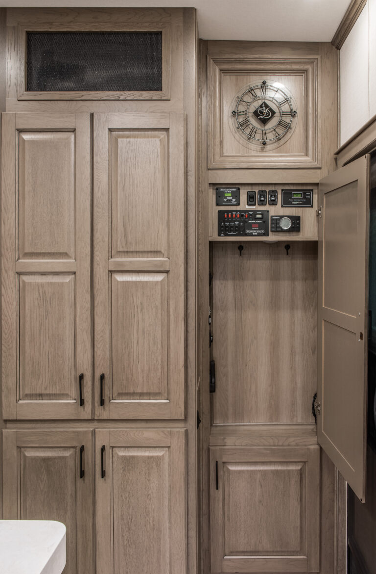 The luxurious wooden cabinets with panelled doors complement a compartment featuring control panels and gauges. A metallic-designed decorative clock elegantly hangs above, hinting at the sophistication of a luxury 5th wheel RV kitchen space.