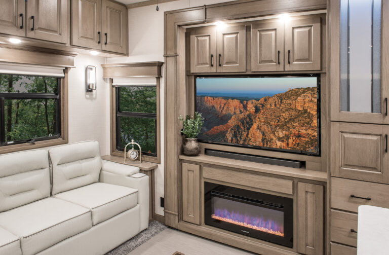 A cozy RV living area with light wood cabinets and a beige sofa exemplifies the charm of luxury RVs. A flat-screen TV displays a canyon landscape above an electric fireplace. Windows reveal green trees, while a small round clock rests on the sofa arm, offering a tranquil retreat.