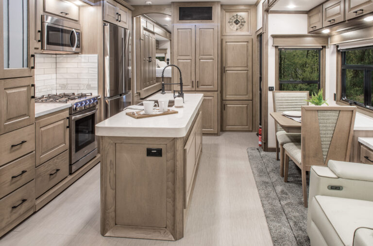 Experience the spacious interior of this luxury RV, complete with a modern kitchen featuring light wood cabinets, a large island with a sink, stainless steel appliances, and a stove. Enjoy cozy dining near a window with lush views. Perfect for those seeking luxury RVs for sale.