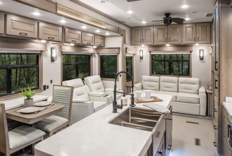 Experience the allure of luxury RVs with this modern interior. Beige furnishings complement a spacious kitchen with a countertop sink, while white sofas and a dining table for two enhance the lounge area. Large windows offer serene forest views, complete with overhead cabinets and a ceiling fan.