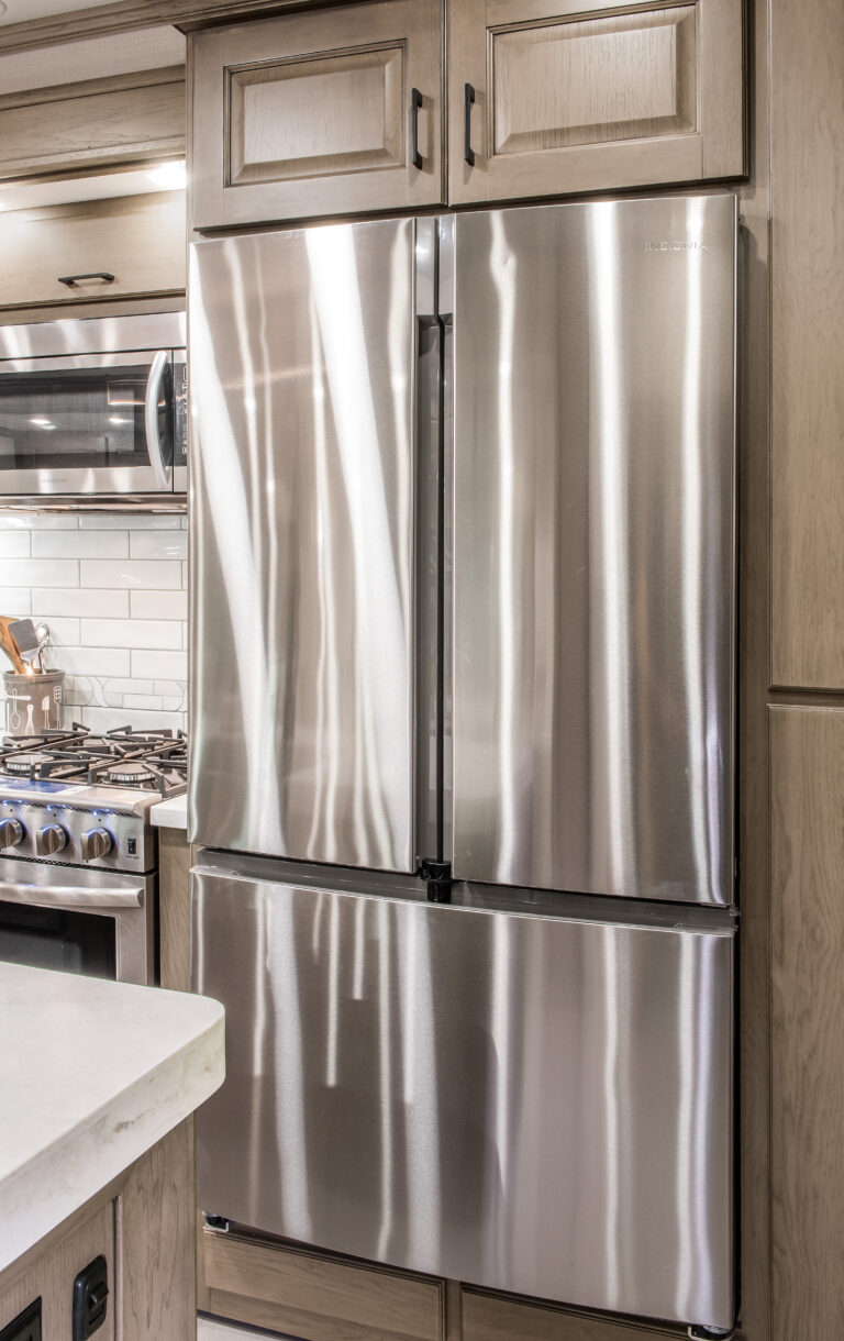 A modern kitchen in a luxury RV boasts a stainless steel French door refrigerator with a lower freezer drawer. The stove and countertop peek out on the left, bordered by white subway tile backsplash, complemented by essential kitchen utensils for culinary convenience.