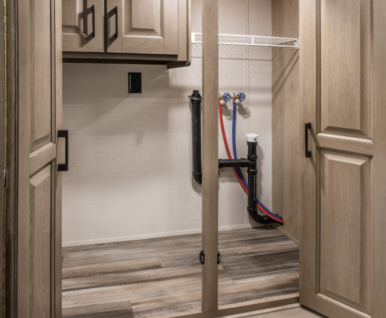 A laundry closet with light wooden cabinets and shelves sits tucked away in a luxury RV. Visible are water hookups and drain pipes against a beige wall, complemented by a wood-patterned floor. Closed cabinet doors complete this elegant yet functional space on the road.