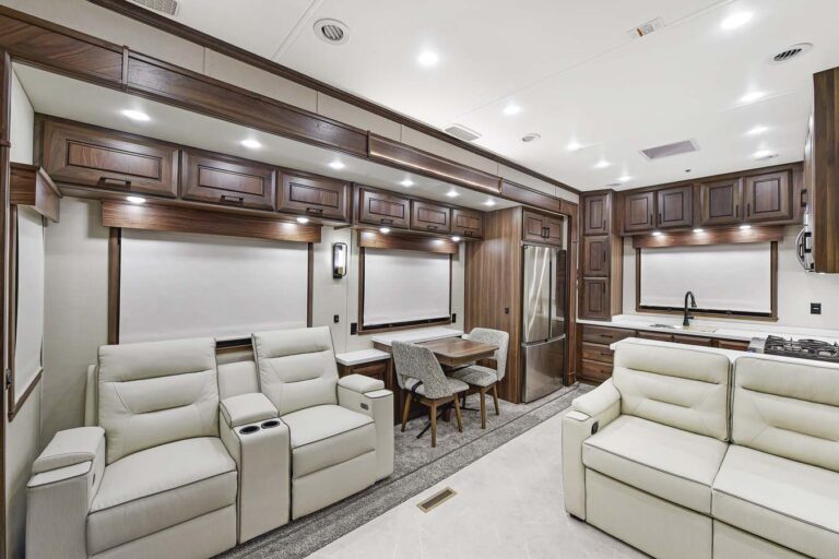 Discover the epitome of luxury RVs with this spacious modern interior. Revel in the cozy seating area featuring light-colored recliners, a chic dining space, and a kitchen boasting a stainless steel fridge. The dark wood cabinets offer contrast and abundant storage in this bright, well-lit haven.