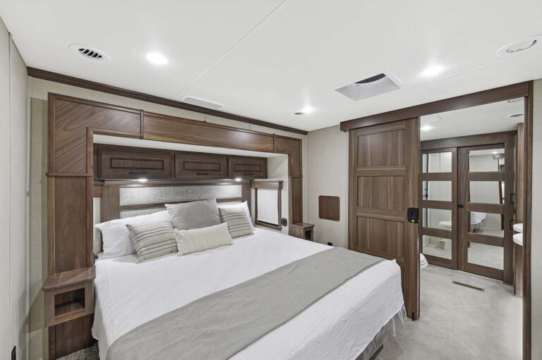 Modern bedroom with a large bed, white and gray bedding, and wood accent wall with storage cabinets. On the right, a sliding wooden door leads to a well-lit bathroom with mirrors and a shower. Neutral walls and recessed lighting evoke the refined elegance of luxury RVs.