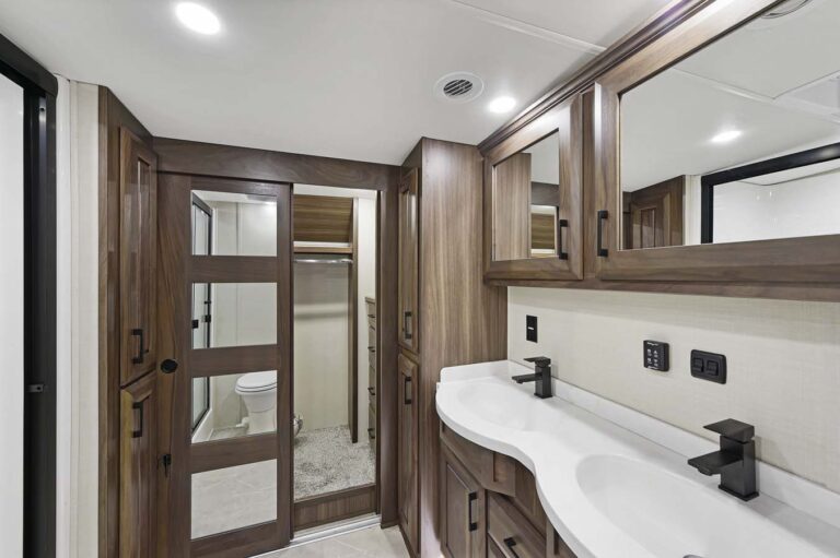 Modern bathroom in luxury RVs features a double sink and wooden cabinetry. On the left, a sliding door reveals a toilet and shower. Walls are light-colored, with ample ceiling lights illuminating the space. Mirrors and black faucets add contrast to this mobile haven.