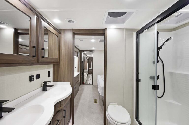 Modern bathroom interior in this luxury RV features wooden cabinets and a double sink vanity on the left. A glass-enclosed shower with a black showerhead is on the right, next to the toilet. A hallway leads to another room, showcasing sophisticated design elements throughout.