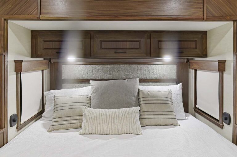Experience the epitome of comfort in this luxury RV. Featuring a bed with crisp white sheets and textured, striped pillows, it promises restful nights. The wooden headboard offers overhead cabinets and adjustable reading lights, while window blinds on either side ensure your privacy.