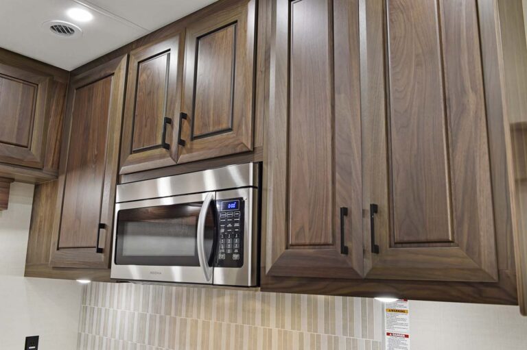 Close-up of a kitchen in luxury RVs with dark wooden cabinets and a built-in stainless steel microwave. The wall beneath features vertically striped tiles in neutral tones, while recessed lighting elegantly illuminates the space.
