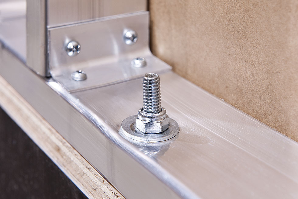 Close-up of a metal bolt secured with a washer and nut on a shiny, reflective surface. The bolt is part of a metallic framework connected by screws in the background, resembling a structural support setup.