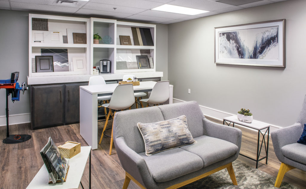 Modern office with grey armchairs, white tables, and framed art. Bookshelves with decor and a coffee setup. Wood flooring and soft lighting create a cozy atmosphere.