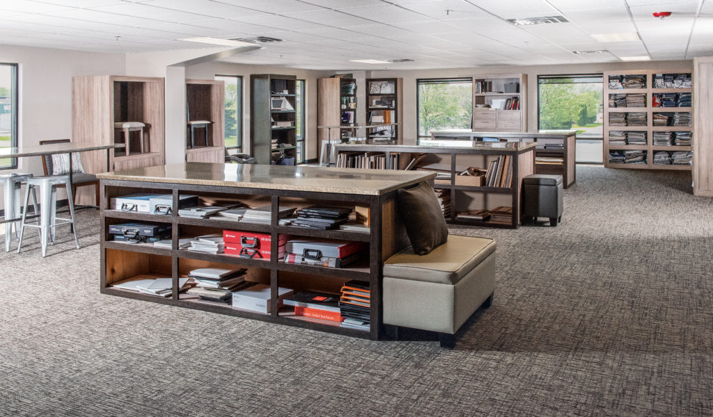 Spacious library with large windows. Bookshelves filled with books and materials line the walls. Two central islands with books on top and seating at one end. Bright natural light creates a welcoming ambiance.