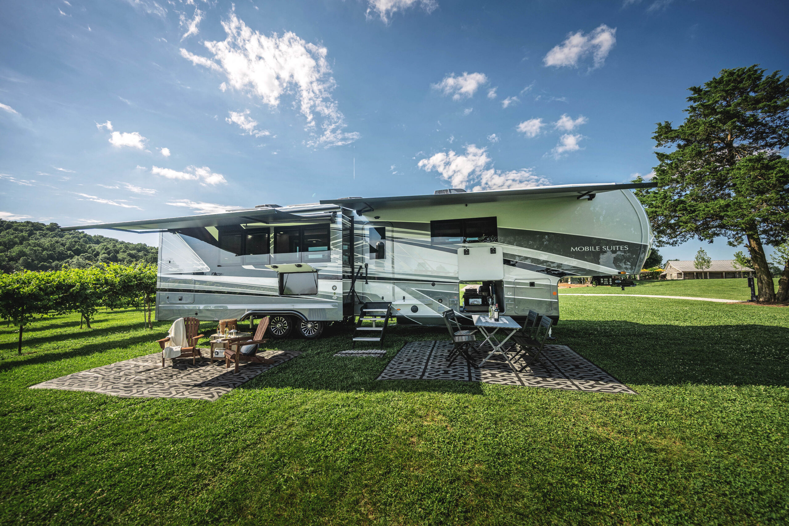 A luxury RV is parked on grass with its awning extended. Outdoor chairs, a table, and rugs are set up on the lawn under a sunny, partly cloudy sky, with trees and distant hills in the background.